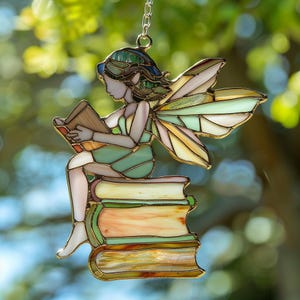 May include: Stained glass fairy reading a book, with green, gold, and white accents. The fairy is seated on a stack of books, with detailed wings and a delicate chain for hanging. A whimsical, decorative piece.