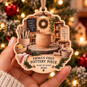 May include: A scalloped wooden ornament featuring a printed pottery studio scene with a central mug on a wheel, tools, and text reading "Emma's First Pottery Piece Made By Hand 2026" with a heart icon, plus signs reading "Pottery People Happier People" and "Good Clay Brighter Days."