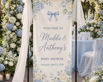 May include: A vertical banner for a baby shower featuring a cream background with blue hydrangea and white rose illustrations. The text reads Welcome to Maddie & Anthony's Baby Shower Saturday, April 11, 2026. A blue bow is centered at the top.