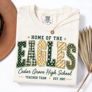 May include: A cream-coloured t-shirt featuring the text HOME OF THE EAGLES in large block letters with varied patterns including polka dots, plaid, checkers, stars, stripes, and leopard print, with Cedar Grove High School Teacher Team Est. 1987.