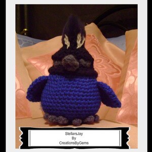 Può includere: Un uccello di peluche lavorato a uncinetto blu e nero con accenti bianchi. L'uccello è seduto su una superficie con motivo rosa e bianco. "StellarsJay By CreationsByGems" è scritto su uno stendardo nero sotto l'uccello.