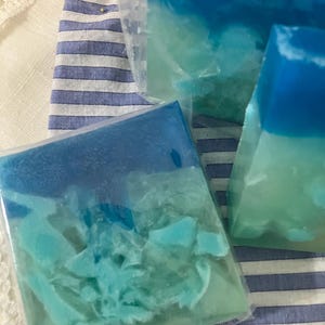 May include: Several handmade soaps in clear packaging. The soaps have a layered design with shades of blue and turquoise, resembling ocean waves. The soaps are displayed on a blue and white striped fabric.
