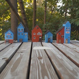 Może przedstawiać: Kolekcja pięciu kolorowych, miniaturowych drewnianych domk&oacute;w. Każdy domek ma unikalny design z malowanymi detalami, takimi jak okna i drzwi. Domki są pomalowane w odcieniach niebieskiego, czerwonego i brązowego i ustawione na drewnianej powierzchni.