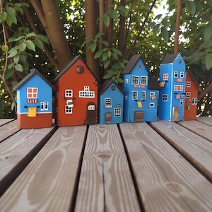Może przedstawiać: Rząd kolorowych, miniaturowych drewnianych domk&oacute;w. Domy pomalowane są w odcieniach niebieskiego, czerwonego i brązowego, z białymi detalami okien i drzwi. Niekt&oacute;re mają małe flagi lub ręczniki. Ułożone są na drewnianej powierzchni.