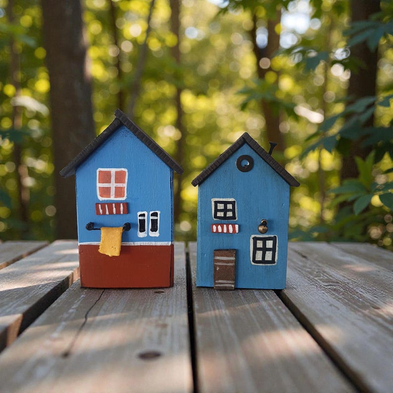 Może przedstawiać: Dwa miniaturowe drewniane domki pomalowane na niebiesko, czerwono i brązowo, z małymi oknami i drzwiami. Jeden ma ż&oacute;łty ręcznik wiszący na drążku. Domki ustawione są na drewnianej powierzchni z rozmytym zielonym tłem.