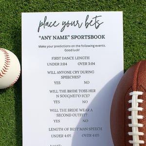 May include: A white sheet with the text "place your bets" and a sports betting game for a wedding. A baseball and a football are on either side of the sheet, which is on a green surface.