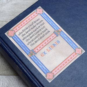 Pode incluir: Um livro com um ex libris decorativo que diz "who folds a leaf down the devil make brown who makes mark or blot the devil toasts hot who steals this book the devil shall cook EX LIBRIS".