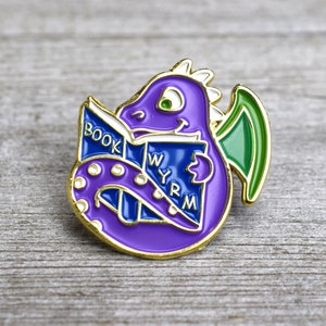 Pode incluir: Um broche de esmalte roxo com um dragão de desenho animado lendo um livro. O dragão tem olhos verdes e uma asa verde. O livro diz "BOOK WYRM" em letras brancas sobre um fundo azul. O broche tem uma borda dourada.