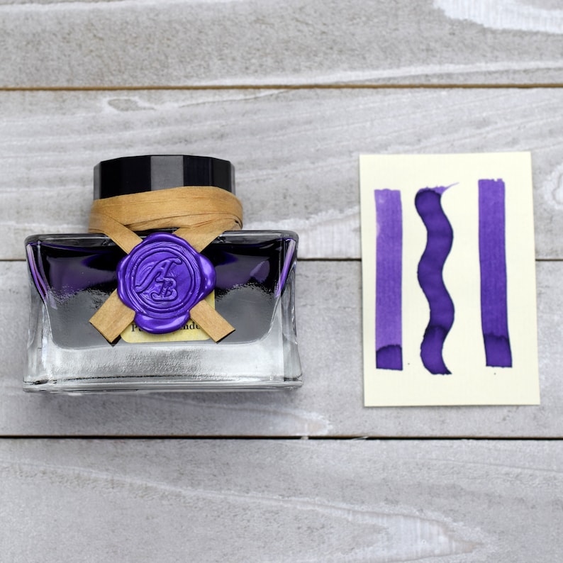 May include: A glass bottle of purple ink with a wax seal and a brown paper label. The bottle is sitting on a light gray wooden surface. A sample of the ink is shown on a white card.