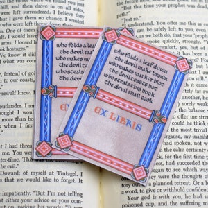Pode incluir: Dois marcadores de livros azuis e vermelhos com uma borda decorativa. O texto no marcador de livros diz "Quem dobra uma folha para baixo, o diabo a torna marrom, quem faz uma marca ou um bloco, o diabo a aquece ao rubro, quem rouba este livro, o diabo o cozinhar&aacute;" e "Ex Libris".