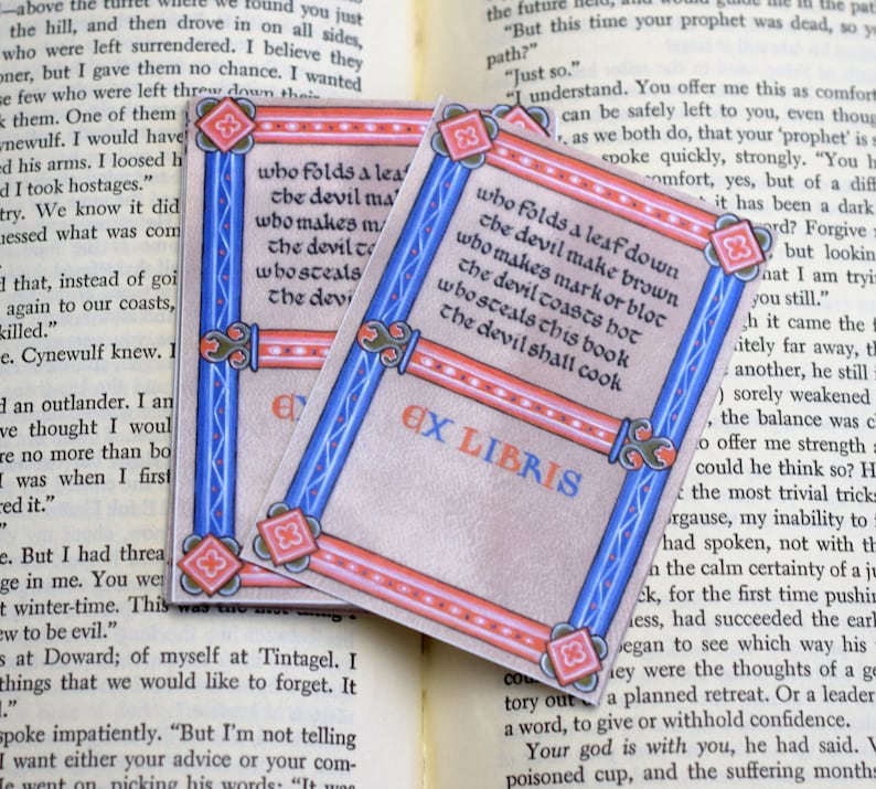 Pode incluir: Dois marcadores de livros azuis e vermelhos com uma borda decorativa. O texto no marcador de livros diz "Quem dobra uma folha para baixo, o diabo a torna marrom, quem faz uma marca ou um bloco, o diabo a aquece ao rubro, quem rouba este livro, o diabo o cozinhar&aacute;" e "Ex Libris".
