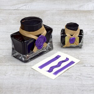 May include: Two glass bottles of purple ink with a wax seal on each. The bottles are on a light wood surface with a purple ink sample card.
