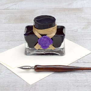 May include: A glass bottle of black ink with a purple wax seal and a brown ribbon. The bottle is sitting on a white surface with a wooden calligraphy pen.