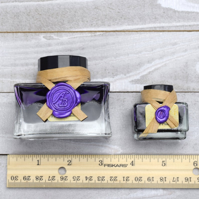 May include: Two glass bottles of purple ink with a wax seal on the top. The bottles are sitting on a wooden surface with a ruler underneath. The ruler is marked with inches.