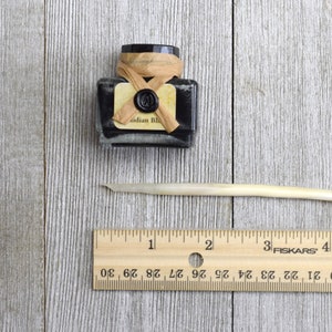 Closeup of a bottle of black writing ink and feather quill next to a ruler for scale