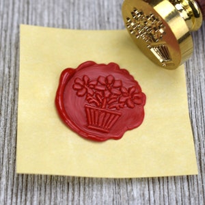 May include: A red wax seal with a design of a potted plant with flowers. The seal is on a light yellow piece of paper.