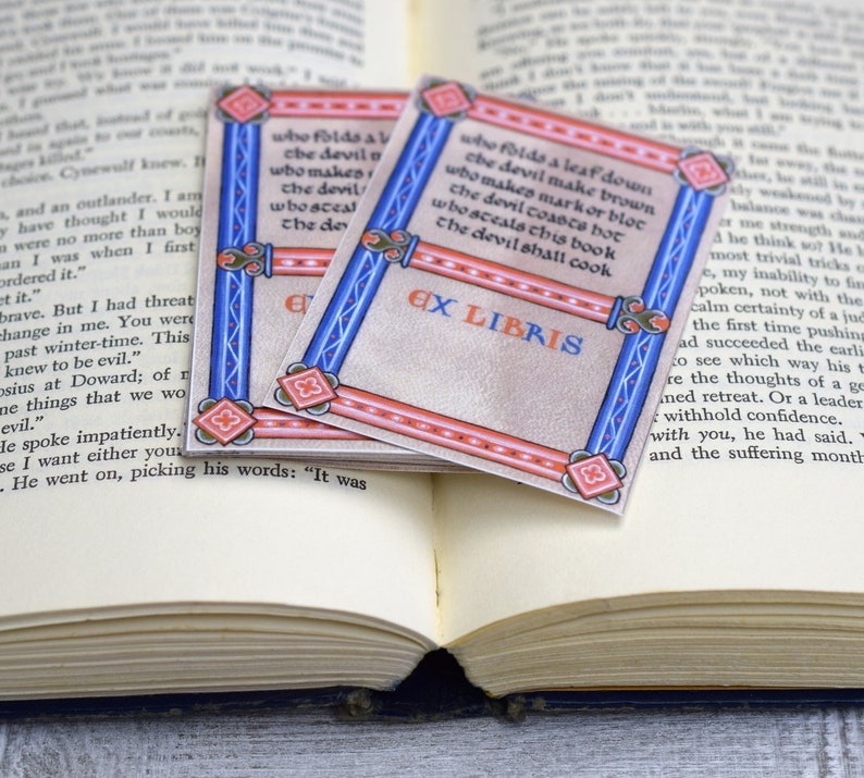 Pode incluir: Dois ex libris decorativos com uma borda vermelha e azul. Os ex libris apresentam um design de estilo medieval com um fundo branco e texto preto. O texto nos ex libris diz "who folds a leaf down the devil make brown who makes markor bloc the devil coasts bot whosceals this book the devil shall cook" e "EX LIBRIS".