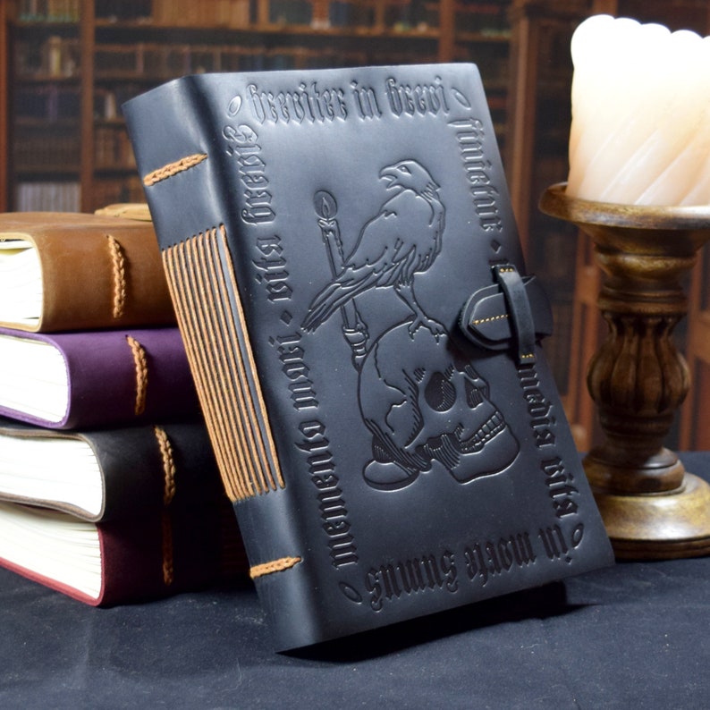 The black leather journal stands upright against a stack of other books. A candle and bookshelves are seen in the background.
