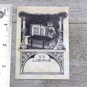 May include: A black and white illustration of an owl perched on a book with two candlesticks on either side. The image is on a beige background with the text "Ex Libris" in a decorative arch.