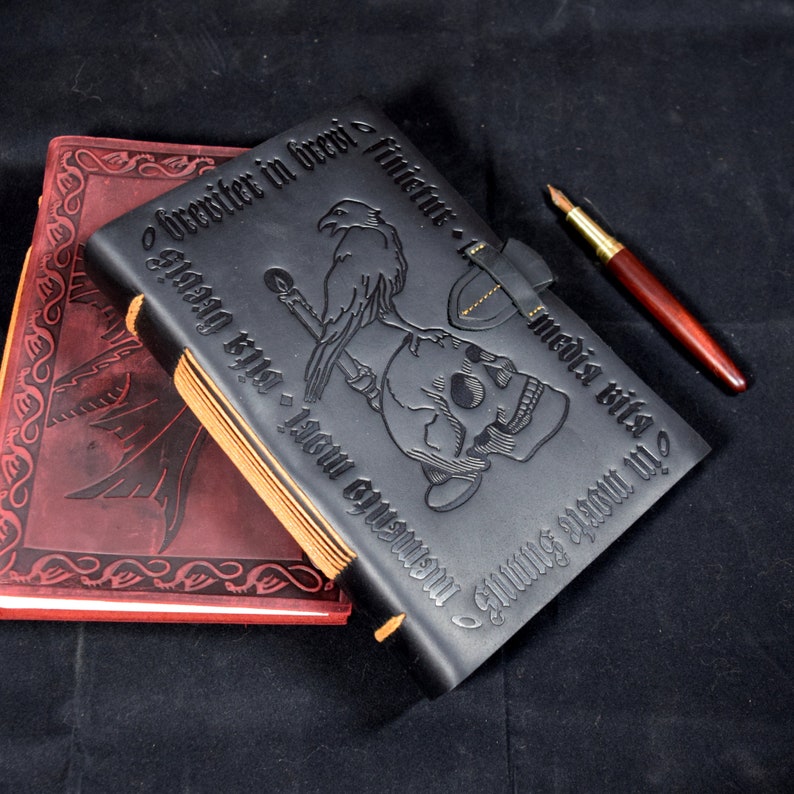 Black leather journal with an embossed Gothic design (featuring skull, raven, candle, and Latin inscription) rests on a black velvet cloth, with a fountain pen and a red journal nearby
