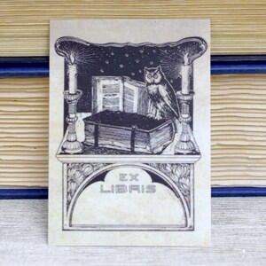 May include: A black and white illustration of an owl perched on a book with candles on either side. The image is framed with a decorative border and includes the text "Ex Libris".