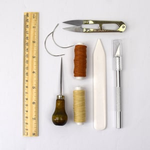 May include: A set of leatherworking tools including a ruler, two needles, two spools of thread, a stitching awl, a bone folder, a craft knife, and a pair of small scissors.