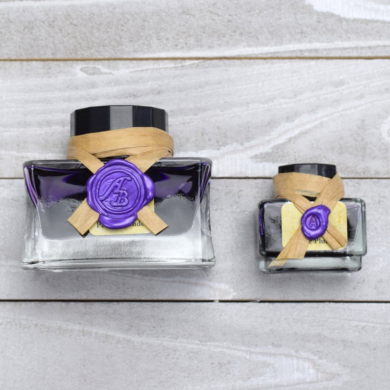 May include: Two glass bottles of purple ink with black caps and brown paper labels. The labels have a purple wax seal with the letter "A" in a circle.