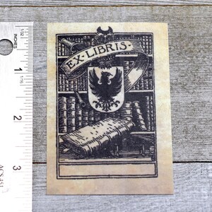 K&ouml;nnte beinhalten: Eine Schwarzwei&szlig;illustration eines Exlibris mit einem Wappen, das einen Vogel mit ausgebreiteten Fl&uuml;geln zeigt. Das Wappen ist von einem Banner mit dem Text "EX-LIBRIS" umgeben und vor einem Hintergrund von B&uuml;cherregalen mit B&uuml;chern platziert. Das Exlibris hat einen leeren Platz f&uuml;r einen Namen oder eine Inschrift.