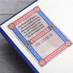 Pode incluir: Um livro com um ex libris decorativo pintado &agrave; m&atilde;o, com uma borda azul e vermelha e uma cita&ccedil;&atilde;o em escrita medieval: "Quem dobra uma folha para baixo, o diabo a torna marrom, quem faz uma marca ou uma mancha, o diabo a torra quente, quem rouba este livro, o diabo o cozinhar&aacute;." O ex libris tamb&eacute;m inclui o texto "Ex Libris".