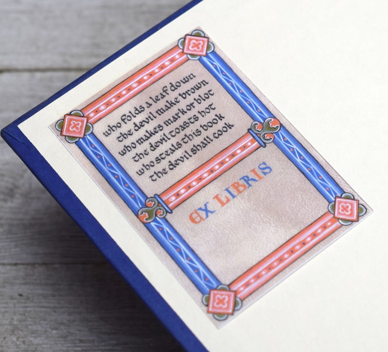 Pode incluir: Um livro com um ex libris decorativo pintado &agrave; m&atilde;o, com uma borda azul e vermelha e uma cita&ccedil;&atilde;o em escrita medieval: "Quem dobra uma folha para baixo, o diabo a torna marrom, quem faz uma marca ou uma mancha, o diabo a torra quente, quem rouba este livro, o diabo o cozinhar&aacute;." O ex libris tamb&eacute;m inclui o texto "Ex Libris".