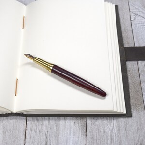 The leather journal is open, revealing hand-sewn blank pages. A fountain pen rests on top of the blank page.
