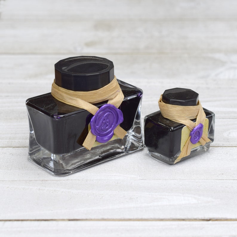 May include: Two glass ink bottles with black lids and purple wax seals. The bottles are filled with dark ink and have a brown ribbon tied around them.