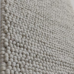 May include: A light grey rug with a textured surface. The rug is covered in small, round, raised bumps, creating a unique tactile experience. The edges of the rug have a woven border. The rug is a neutral colour, suitable for various interior design styles.