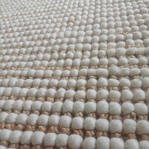 Puede incluir: Primer plano de una alfombra texturizada con un patrón repetitivo. La alfombra presenta filas de mechones blancos y redondeados que se alternan con bandas de material tejido de color natural. El diseño general crea una superficie táctil y visualmente atractiva.