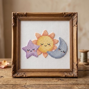 May include: A framed cross-stitch artwork featuring a smiling sun, moon, and star. The sun is yellow and orange, the star is purple, and the moon is blue. The frame is a dark wood with ornate detailing. The artwork is displayed on a wooden surface.