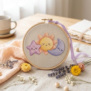 May include: A cross-stitch embroidery hoop featuring a smiling sun, star, and moon design. The sun is yellow and orange, the star is purple, and the moon is blue. The hoop is made of wood and has a lavender ribbon attached.