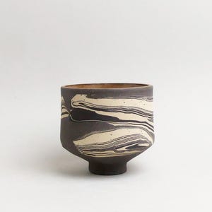 May include: A small ceramic cup featuring a marbled pattern of dark charcoal gray and off-white swirls. The vessel has a cylindrical body with a slightly tapered base and a smooth, matte finish, showcasing a distinct layered clay aesthetic.