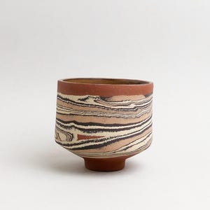 May include: A small ceramic cup featuring a marbled pattern of terracotta, cream, and dark brown horizontal swirls. The vessel has a cylindrical body with a slightly tapered base and a smooth, matte finish, showcasing a distinct layered clay design.