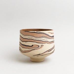 May include: A ceramic cup featuring a marbled pattern of cream, tan, and dark brown swirling lines. The vessel has a rounded body that tapers down to a small circular base, with a smooth, open top rim and a visible interior surface.