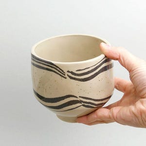 Könnte beinhalten: Eine beige Keramikschale mit dunklen, wellenförmigen Linien am oberen Rand. Die Schale hat eine gesprenkelte Textur und eine abgerundete Form. Der Innenraum ist hellbeige. Die Schale wird von einer Hand gehalten.