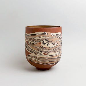 Könnte beinhalten: Eine handgefertigte Keramiktasse mit einem einzigartigen Marmormuster. Die Tasse hat eine rotbraune Außenseite mit wirbelnden Mustern in Creme-, Beige- und Dunkelbrauntönen. Sie hat eine abgerundete Form und einen kleinen, flachen Boden. Der Innenraum ist dunkelbraun.