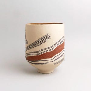 Op de afbeelding: Een crèmekleurige keramische beker met een brede opening en een licht taps toelopende basis. De beker is versierd met horizontale banden van donkerbruine, zwarte en roodbruine strepen. De binnenrand heeft een lichtbruine glazuur.