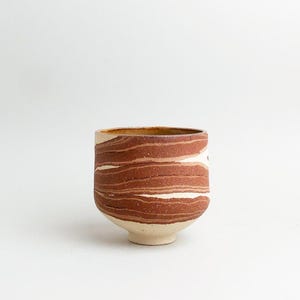 Könnte beinhalten: Eine handgefertigte Keramiktasse mit einem einzigartigen Design. Die Tasse weist horizontale Bänder aus rotbraunem und cremefarbenem Ton auf. Sie hat einen abgerundeten Körper und einen kleinen, fußartigen Sockel. Der Rand ist dunkler braun.