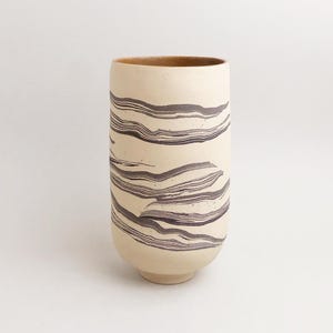 Könnte beinhalten: Eine zylindrische Keramikvase mit einer matten, cremefarbenen Außenseite, die horizontale, wellenförmige, dunkelbraune Marmormuster aufweist. Das Innere der Vase ist glatt und einfarbig hellbraun, der Boden ist leicht verjüngt mit einem kleinen Fuß.