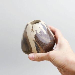 Könnte beinhalten: Eine kleine, handgefertigte Keramikvase mit einer einzigartigen, organischen Form. Die Vase hat ein marmoriertes Design in Braun- und Cremetönen, das eine natürliche, erdige Ästhetik erzeugt. Die Vase wird in einer Hand gehalten, um ihre Größe und Form zu zeigen.