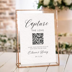以下が含まれることがあります： 「Capture the Love」の文字とQRコードが書かれた額縁入りのサイン。サインには「Olivia & Henry」と日付「04.12.26」も含まれています。フレームはローズゴールド色で、背景はウェディングケーキのぼやけた画像です。
