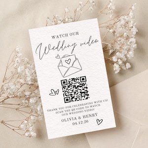 May include: A white wedding invitation card with the text "Watch Our Wedding Video" in elegant script. It features a QR code, an envelope with a heart, and the names Olivia & Henry with the date 04.12.26. The card is decorated with small hearts.