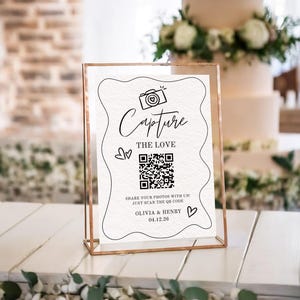 May include: A clear acrylic sign with a gold metal frame. The sign has the words "Capture The Love" with a camera graphic and a QR code. The sign also includes the names Olivia & Henry and the date 04.12.26. A wedding decor item.