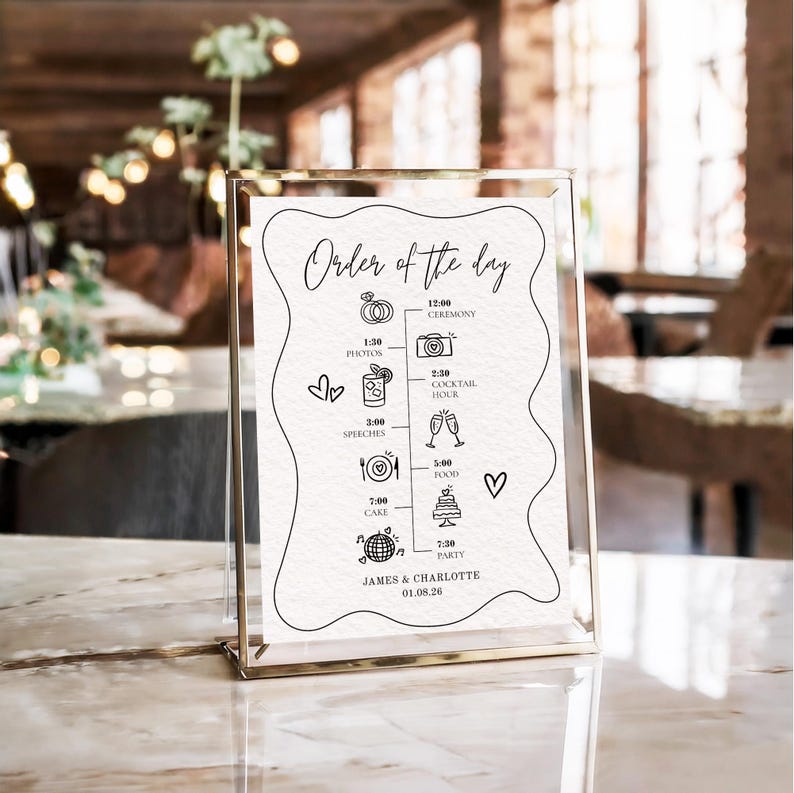 May include: A gold-framed wedding sign displaying the "Order of the Day" in black script. The sign includes icons and times for the ceremony, photos, cocktail hour, speeches, food, cake, and party. The names "James & Charlotte" and the date are at the bottom.