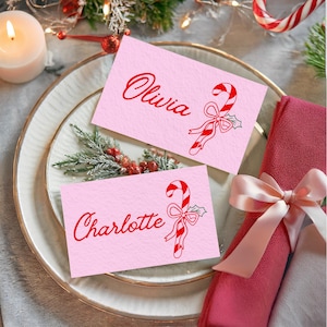Peut inclure: Deux cartons roses avec du texte rouge et des illustrations de cannes en sucre. Une carte indique "Olivia", l'autre "Charlotte". Les cartes sont sur une assiette blanche avec une bordure dorée, à côté d'une serviette rouge et d'une bougie allumée.